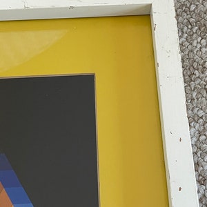 May include: A framed abstract art piece with a white distressed wooden frame. The artwork features a geometric design with a black background, a yellow border, and a colorful section with blue, purple, and brown hues.
