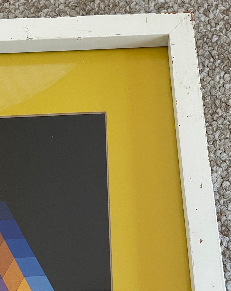 May include: A framed abstract art piece with a white distressed wooden frame. The artwork features a geometric design with a black background, a yellow border, and a colorful section with blue, purple, and brown hues.