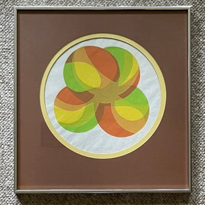 May include: A framed print of an abstract geometric design featuring overlapping circles in shades of orange, yellow, and green. The print is set against a brown background and has a gold-colored frame.