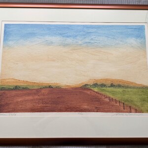 May include: Framed landscape art print featuring a field with a red-brown foreground, green grass, and a fence. The sky transitions from blue to beige. The print is in a brown frame with a white mat.