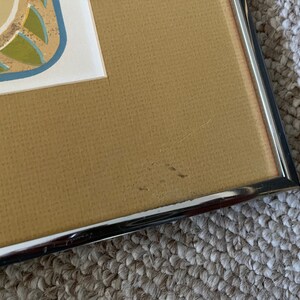 May include: Close-up of the bottom right corner of a framed picture. The frame is silver metal and the mat is a light brown color. There are a few small scratches on the mat.
