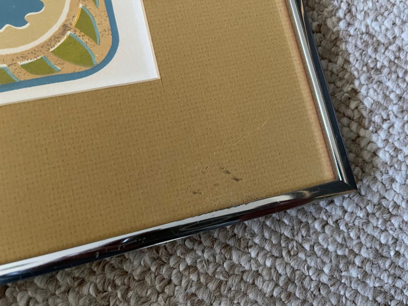 May include: Close-up of the bottom right corner of a framed picture. The frame is silver metal and the mat is a light brown color. There are a few small scratches on the mat.