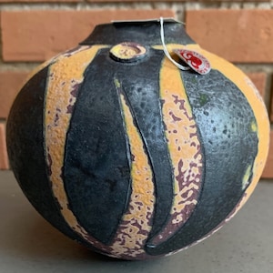 May include: A small, round ceramic vase with a black, yellow, and purple glaze. The vase has a textured surface and a unique, abstract design.