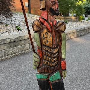May include: Hand-carved wooden statue of a figure with a hat and staff. The statue is painted with red, green, and yellow accents. The figure is standing on a wooden base and holding a staff with a fleur-de-lis design.