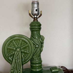 May include: Green ceramic lamp with a decorative wheel design. The lamp has a textured, carved appearance and a standard light bulb. The lamp is on a wooden surface.
