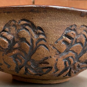 May include: A handmade, brown ceramic bowl with a dark brown rim and intricate floral carvings. The bowl's exterior features a textured surface and a rounded shape, perfect for serving or display.