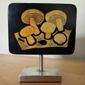 May include: A black rectangular plaque with a stand, featuring a stylized image of mushrooms in shades of yellow and brown. The plaque has a textured surface and is decorated with small black insects.