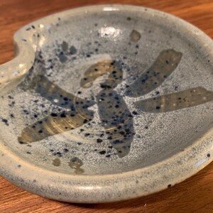May include: A small, light blue ceramic dish with a white glaze and a speckled pattern. The dish has a unique shape with a curved edge and a small indentation in the center. The dish is decorated with a stylized design of a flower or star in a light brown color.