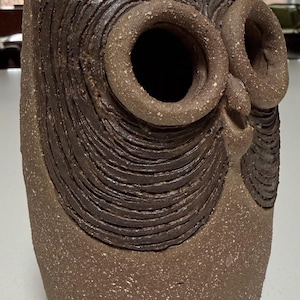 May include: A brown ceramic owl sculpture with a textured surface. The owl has large, circular eyes and a detailed, layered design around the eyes and head. The sculpture is a decorative item.