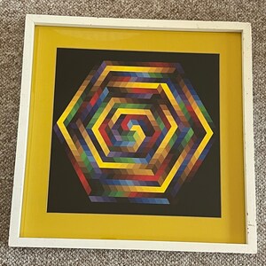 May include: Geometric art print in a white frame with a yellow mat. The artwork features a colorful, spiraling hexagonal design on a black background. The colors include shades of red, yellow, blue, green, and brown. The frame is rectangular.