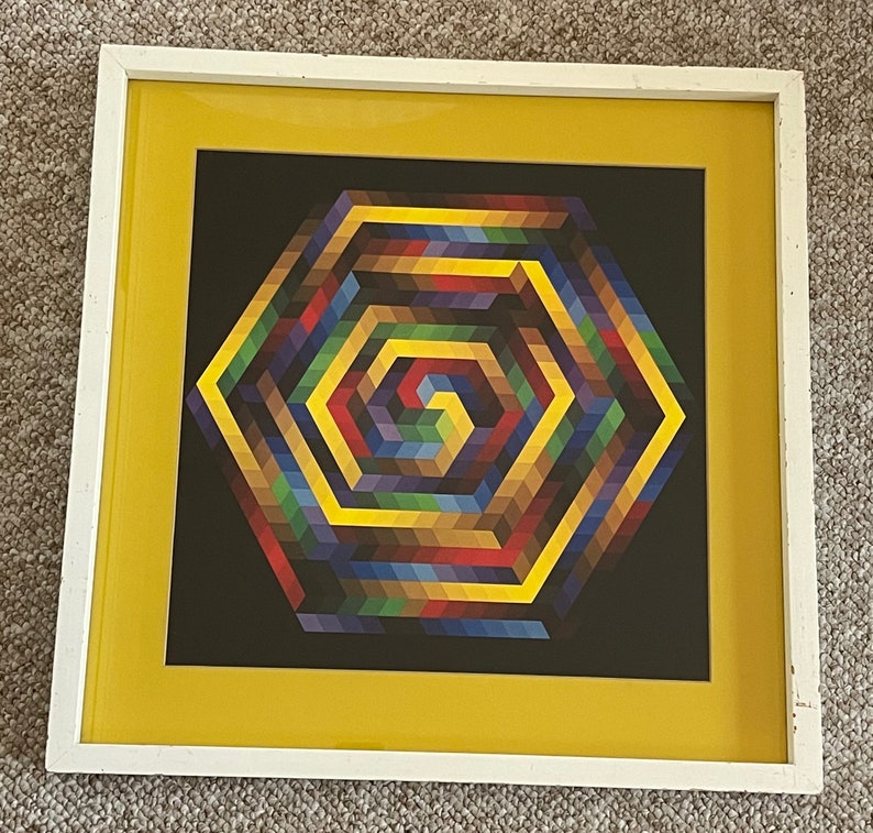 May include: Geometric art print in a white frame with a yellow mat. The artwork features a colorful, spiraling hexagonal design on a black background. The colors include shades of red, yellow, blue, green, and brown. The frame is rectangular.