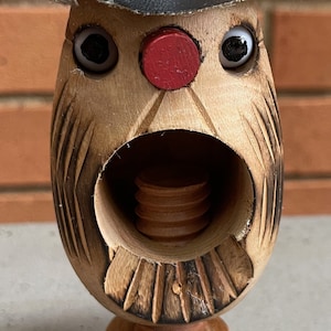 May include: Wooden nutcracker with a carved face, wearing a black hat with a brown brim. The nutcracker has a red nose and large, white eyes.