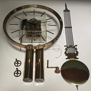 May include: A gold-toned clock kit with a clear face and black hands. The kit includes two weights, a pendulum, a winding key, and a decorative top piece. The clock face has the letter 'H' at the bottom.