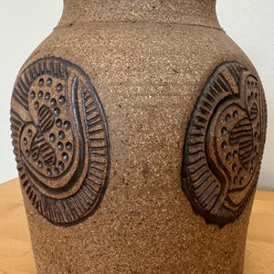 May include: A brown ceramic vase with a textured surface and two embossed designs. The designs are in the shape of a heart with a swirl pattern.
