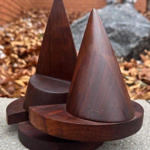 May include: Two dark brown wooden sculptures with conical tops and circular bases. The sculptures are made of a rich, dark wood and are displayed on a concrete surface. The background features fallen leaves and a blurred view of a rock.