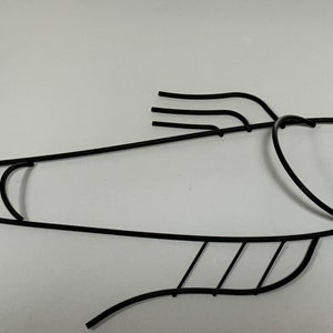 May include: Black metal fish wall decor with a stylized design. The fish has a curved body, a long tail, and a detailed head with a small eye.