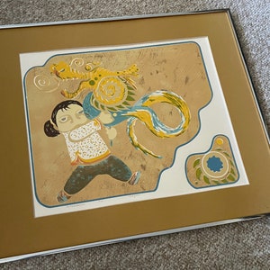 May include: A framed print of a colorful illustration depicting a young person with a dragon. The dragon is yellow, blue, and green, and the person is wearing a white shirt with a blue and yellow pattern. The background is a light brown color.