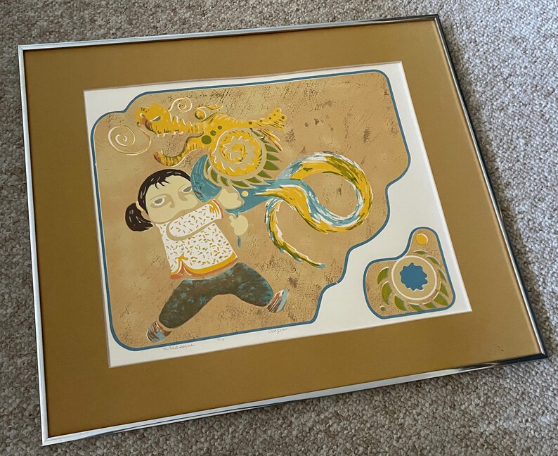May include: A framed print of a colorful illustration depicting a young person with a dragon. The dragon is yellow, blue, and green, and the person is wearing a white shirt with a blue and yellow pattern. The background is a light brown color.