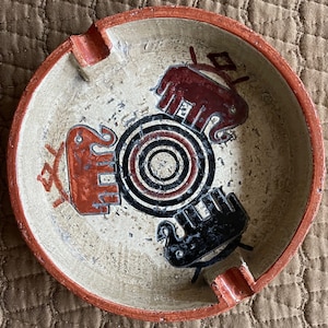 May include: A round, ceramic ashtray with a red rim and a white interior. The ashtray features a black and white concentric circle design in the center, surrounded by three stylized animal figures. The figures are painted in red, black, and white.