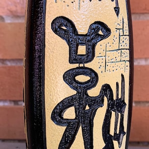 May include: A black and yellow ceramic vase with a stylized design of a person and a brick pattern. The vase has a textured surface and a rounded shape.