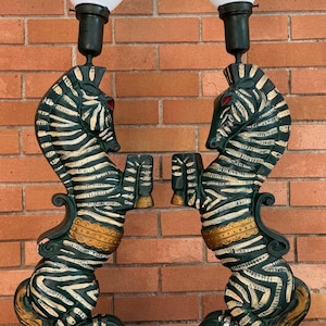 K&ouml;nnte beinhalten: Paar Vintage-Tischlampen mit zebraf&ouml;rmigen Sockeln. Die Lampen haben wei&szlig;-schwarze Zebrastreifen, rote Augen und goldfarbene Akzente. Jede Lampe hat einen wei&szlig;en, konisch geformten Schirm. Die Lampen stehen auf einem gr&uuml;nen Sockel.