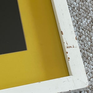 May include: Close-up of a framed artwork with a distressed white wooden frame. The artwork features a black rectangular shape set against a vibrant yellow background. The frame shows signs of wear.