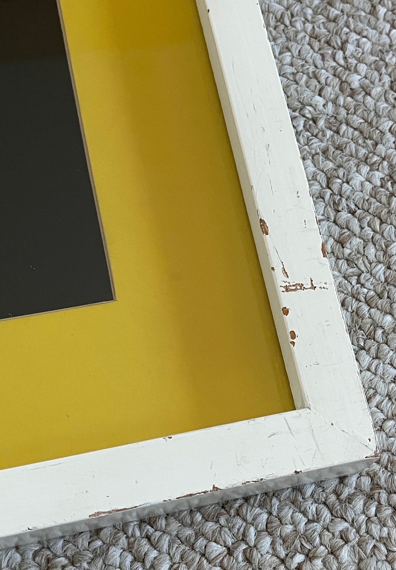 May include: Close-up of a framed artwork with a distressed white wooden frame. The artwork features a black rectangular shape set against a vibrant yellow background. The frame shows signs of wear.