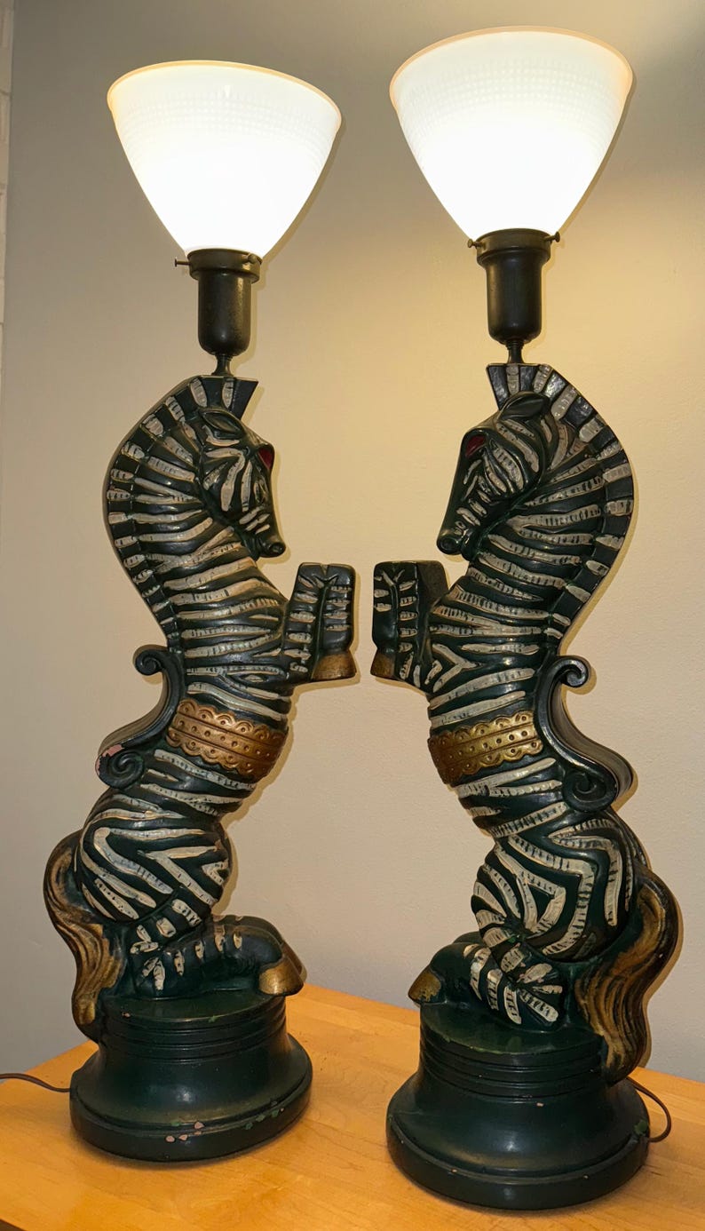 K&ouml;nnte beinhalten: Paar dekorative Tischlampen mit zebraf&ouml;rmigen Sockeln mit schwarz-wei&szlig;en Streifen, goldenen Akzenten und wei&szlig;en Glasschirmen. Die Lampen haben einen dunkelgr&uuml;nen Sockel und sind so gestaltet, dass sie sich gegen&uuml;berstehenden Zebras &auml;hneln.
