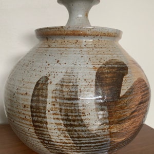 May include: A ceramic jar with a lid and a unique design. The jar is glazed in shades of brown, gray, and white, with a swirling pattern. The lid has a knob on top.