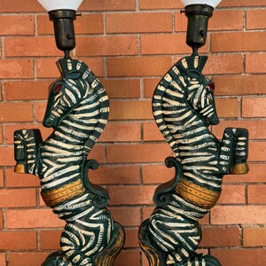 K&ouml;nnte beinhalten: Paar Vintage-Lampen im Zebra-Design. Jede Lampe zeigt eine gr&uuml;n-wei&szlig;e Zebrafigur, die ein kleines Objekt h&auml;lt, mit roten Augen und goldenen Akzenten. Die Lampen haben wei&szlig;e, konisch geformte Schirme und stehen auf einem gr&uuml;nen Sockel.