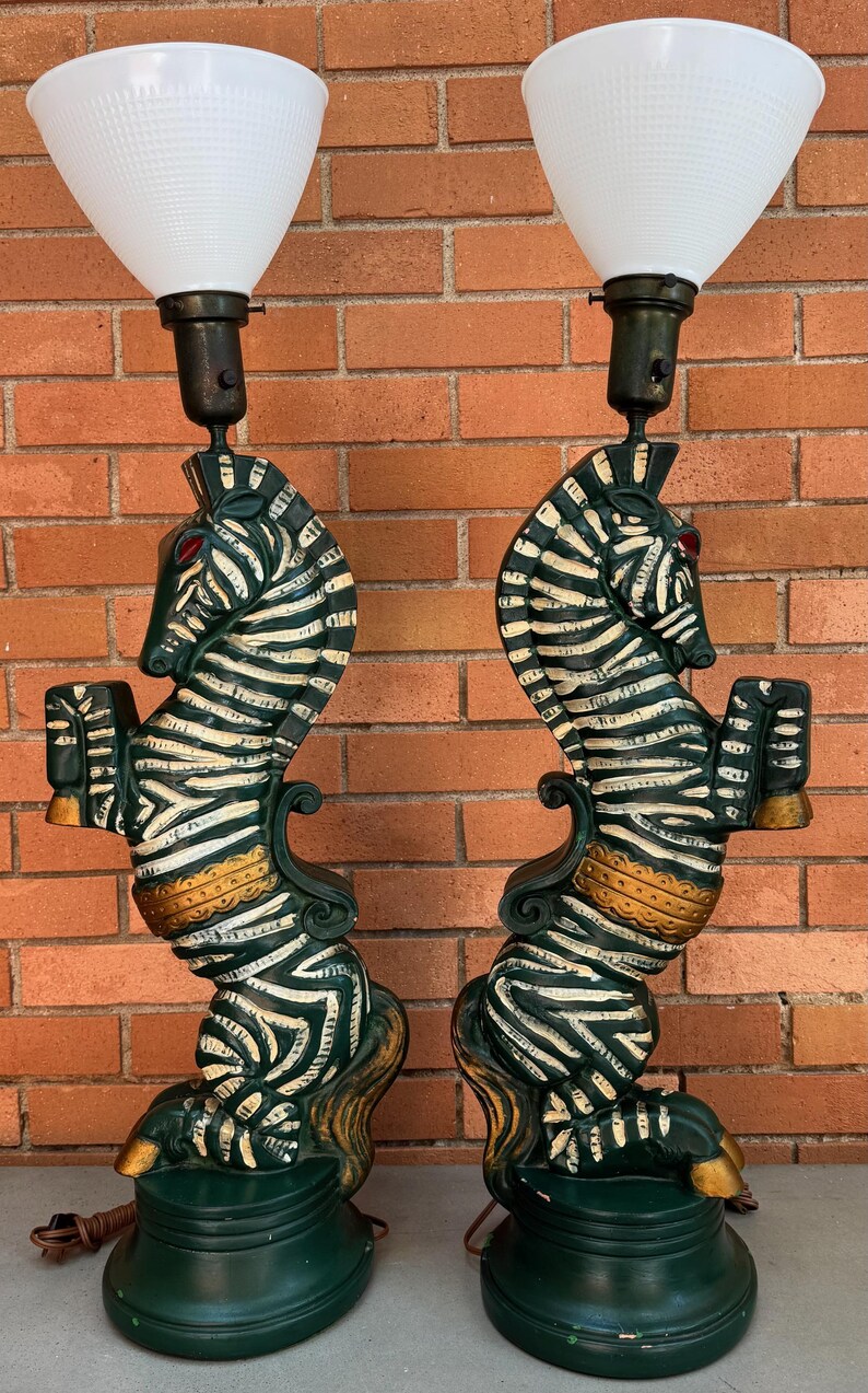 K&ouml;nnte beinhalten: Paar Vintage-Lampen im Zebra-Design. Jede Lampe zeigt eine gr&uuml;n-wei&szlig;e Zebrafigur, die ein kleines Objekt h&auml;lt, mit roten Augen und goldenen Akzenten. Die Lampen haben wei&szlig;e, konisch geformte Schirme und stehen auf einem gr&uuml;nen Sockel.