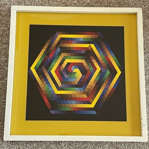 May include: Geometric art print in a white frame with a yellow mat. The artwork features a colorful, hexagonal design with a spiral center. The color palette includes shades of yellow, blue, red, and brown against a black background.