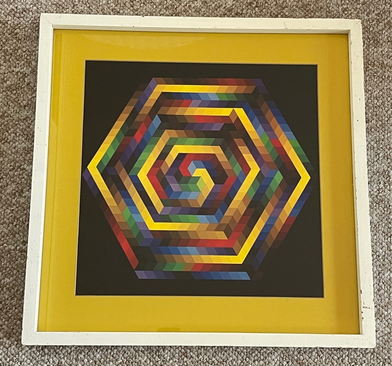 May include: Geometric art print in a white frame with a yellow mat. The artwork features a colorful, hexagonal design with a spiral center. The color palette includes shades of yellow, blue, red, and brown against a black background.