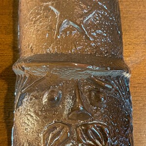 May include: A glass bottle stopper with a textured surface and a face sculpted into the glass. The face has a mustache and the word "Puff" is written on the bottom.