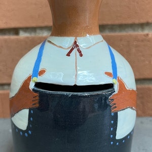 May include: A ceramic piggy bank shaped like a person wearing a white shirt, blue suspenders, and black pants. The piggy bank has a slot for coins in the front.