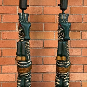K&ouml;nnte beinhalten: Paar dekorative Tischlampen mit Totempfahl-Design. Die Lampen haben einen dunkelgr&uuml;nen Sockel und K&ouml;rper mit goldenen und wei&szlig;en Akzenten. Jede Lampe hat einen wei&szlig;en, konisch geformten Schirm.