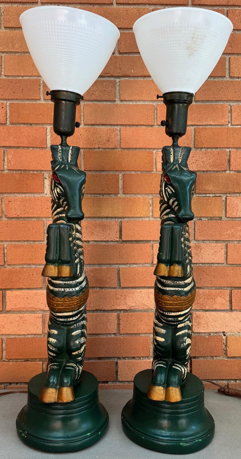 K&ouml;nnte beinhalten: Paar dekorative Tischlampen mit Totempfahl-Design. Die Lampen haben einen dunkelgr&uuml;nen Sockel und K&ouml;rper mit goldenen und wei&szlig;en Akzenten. Jede Lampe hat einen wei&szlig;en, konisch geformten Schirm.