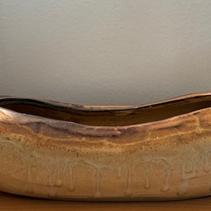 May include: A ceramic planter shaped like a canoe with a light brown glaze and a darker brown glaze inside. The planter is made of a smooth, textured ceramic.