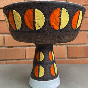 May include: A brown ceramic pedestal bowl with a white base. The bowl is decorated with a pattern of orange and yellow half circles.