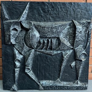 May include: A dark gray, abstract relief sculpture of a bull. The artwork features geometric shapes and textured surfaces, with a rough, carved appearance. The bull's form is stylized, with prominent horns and a detailed mouth.