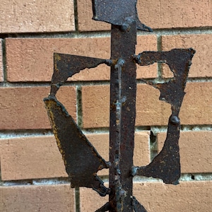 May include: A rusted metal sculpture with a vertical central support and various geometric shapes welded to it. The sculpture has a rough, textured surface and is set against a brick wall.