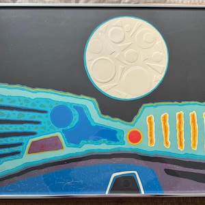 May include: Abstract art print in a silver frame. The artwork features a large white moon with textured details against a dark gray sky. Below, a landscape is depicted with blue, turquoise, yellow, and purple shapes. Signed by the artist.