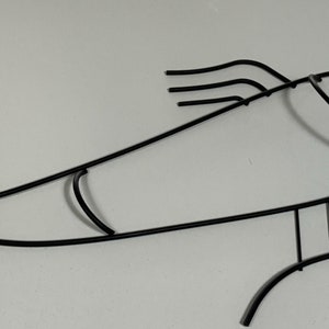 May include: Black metal fish wall decor. The fish is stylized with a curved body and a long, thin tail. The fish's head is rounded with a small mouth and a single eye.