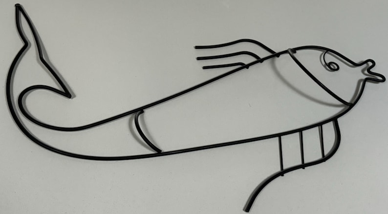 May include: Black metal fish wall decor. The fish is stylized with a curved body and a long, thin tail. The fish's head is rounded with a small mouth and a single eye.