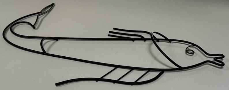 May include: Black metal fish sculpture with a stylized design. The fish is depicted in a swimming position with its tail curved upwards and its fins spread out.