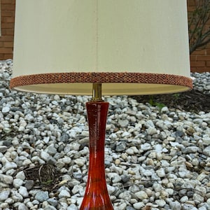 May include: A red ceramic table lamp with a white lampshade and a brown trim. The lamp has a rounded base and a tall, slender neck.