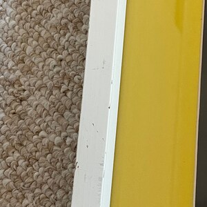 May include: Close-up of a framed artwork with a white wooden frame. The frame surrounds a vibrant yellow background. The frame shows signs of wear, with visible scratches and imperfections. The artwork is placed on a textured, neutral-colored carpet.