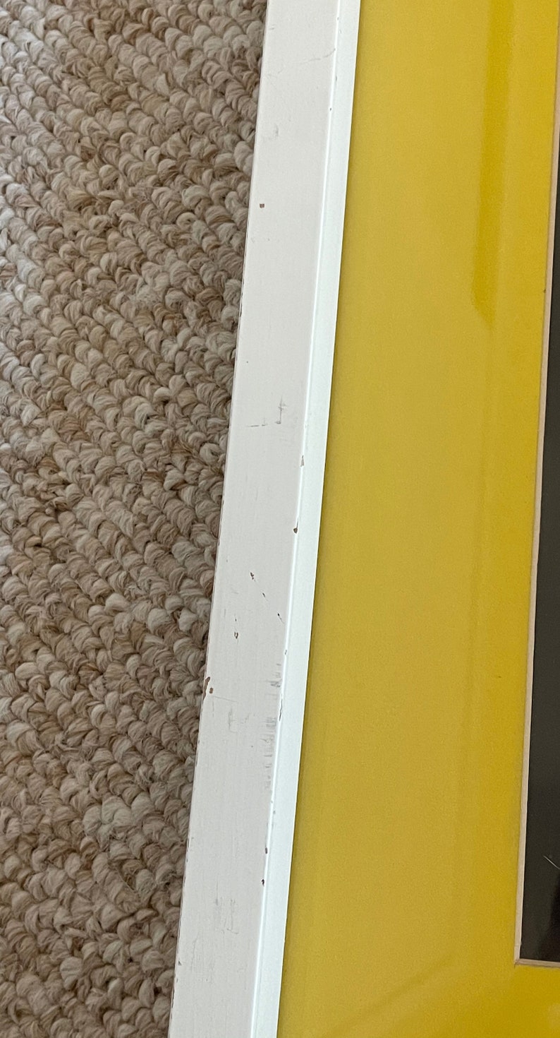 May include: Close-up of a framed artwork with a white wooden frame. The frame surrounds a vibrant yellow background. The frame shows signs of wear, with visible scratches and imperfections. The artwork is placed on a textured, neutral-colored carpet.