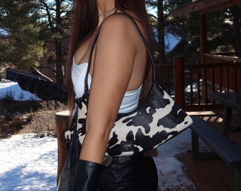 May include: A shoulder bag featuring a black and white cow print pattern with a smooth texture. The bag has a curved silhouette, a single black strap, and a small rectangular black label with gold lettering on the lower front section.