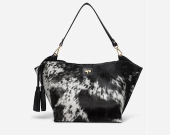 May include: A black and white cowhide shoulder bag featuring a textured animal print pattern, a single black leather strap with gold-tone hardware, and a decorative black leather tassel hanging from the side. A small black label displays the text Lypa.